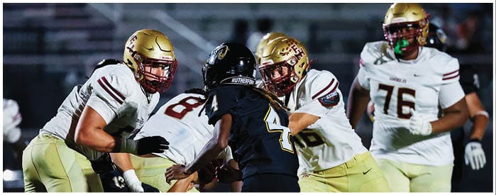 People wearing football helmets and uniforms are engaged in a game on a field with empty bleachers in the background. Description generated by AI