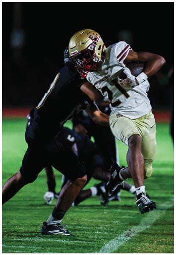 A person wearing a gold football helmet and white jersey with the number 21 is running with a football while being tackled by another player in a dark uniform. Description generated by AI