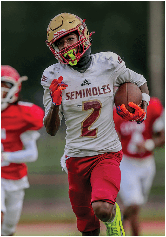 A person wearing a football helmet and a white jersey with the word “Seminoles" and the number 2 is running on a field with another player in red blurred in the background. Description generated by AI