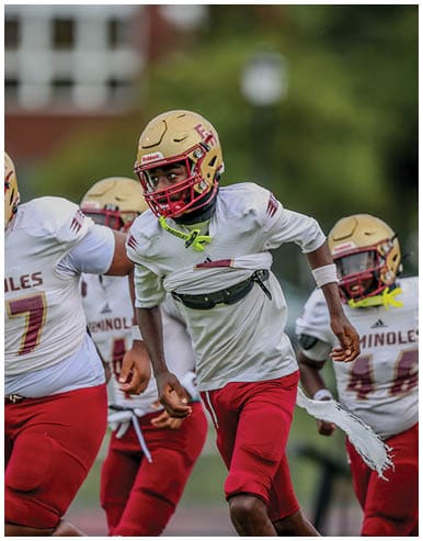 People wearing football helmets and uniforms are running on a field during a game or practice. Description generated by AI