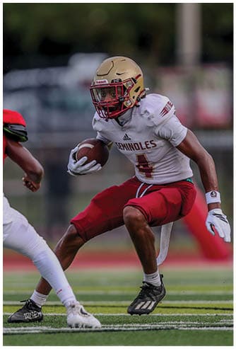 A person wearing a football uniform with a gold helmet and red pants is running with a football on a field while another person in white pants and cleats approaches. Description generated by AI