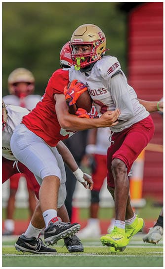 A person wearing a white and maroon football uniform with bright yellow cleats is running while holding a football and being tackled by two people in red and white uniforms on a green field. Description generated by AI