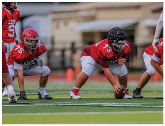 A person wearing a red sports jersey and protective helmet is crouched on a football field holding a football, preparing to snap it to a teammate. Description generated by AI