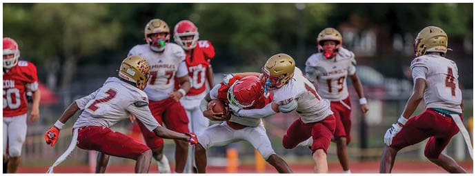 A group of people wearing football helmets and uniforms are actively playing on a field with one person holding the ball while others attempt to tackle or block. Description generated by AI