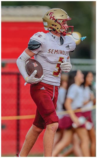 A person wearing a football uniform with a helmet and holding a football is walking on a field with blurred people in the background. Description generated by AI