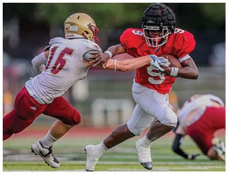 Two people in football uniforms are engaged in a play on a field, with one person in a red jersey running while holding the ball and the other in a white jersey attempting to tackle them. Description generated by AI