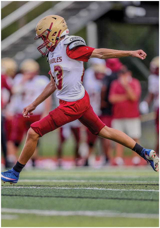 A person wearing a football uniform with a gold helmet and red pants is in mid-kick on a football field. Description generated by AI