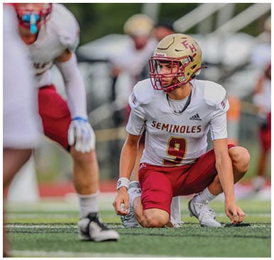 A person wearing a football helmet and uniform with the word Seminoles and the number 9 is kneeling on a football field while other players stand nearby. Description generated by AI