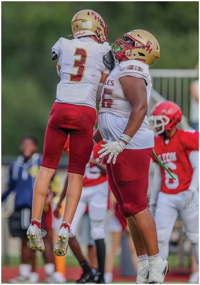 A person wearing a white and red football uniform with the number three is jumping in the air during a game while other players and cheerleaders are visible in the background. Description generated by AI