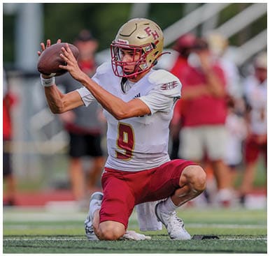 A person wearing a football uniform with a helmet is kneeling on one knee while holding a football in both hands, preparing to throw it on a field. Description generated by AI