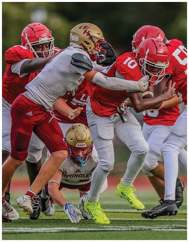 People in football uniforms are actively engaged in a play on a grassy field with one player in red holding the ball while others in red and white attempt to block or tackle. Description generated by AI