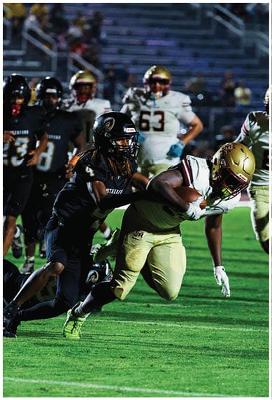 A group of people in football uniforms are actively playing on a field with one person in dark clothing tackling another person in light clothing who is holding a football. Description generated by AI