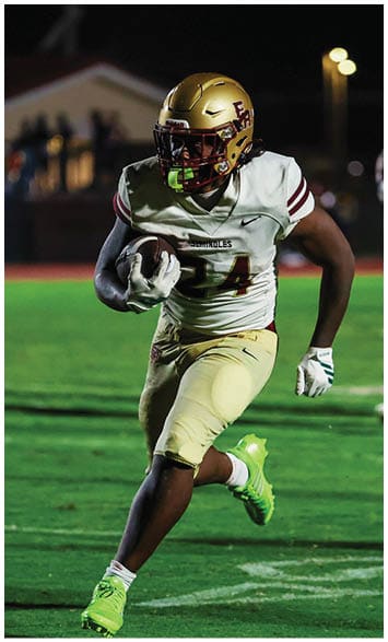 An adult wearing a football uniform with neon green cleats is running on a grass field while holding a football. Description generated by AI