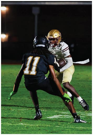 Two people in football uniforms are engaged in a play on a grassy field at night, with one person holding the ball and the other preparing to defend. Description generated by AI