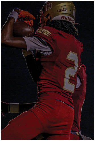 A person wearing a football helmet, white jersey, red shorts, and neon yellow cleats is crouched on a football field holding a football. Description generated by AI