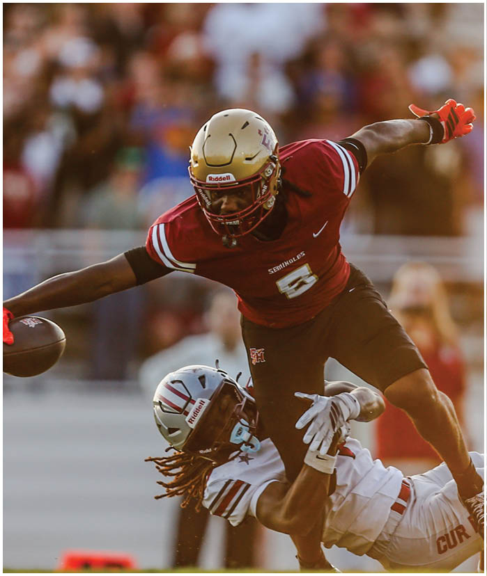 A person in a dark uniform is running with a football while being tackled by a person in a white and gold uniform during a football game. Description generated by AI