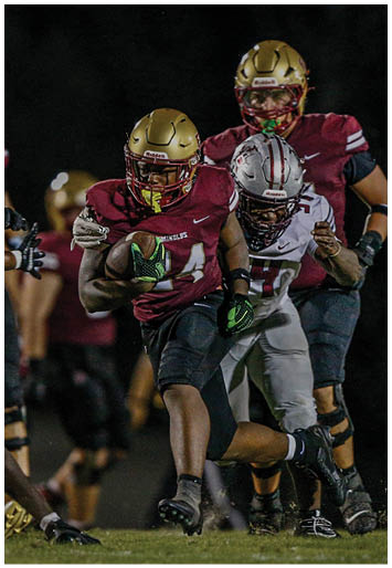 Two people wearing gold football helmets and white jerseys are engaged in a play on a dimly lit field at night. Description generated by AI
