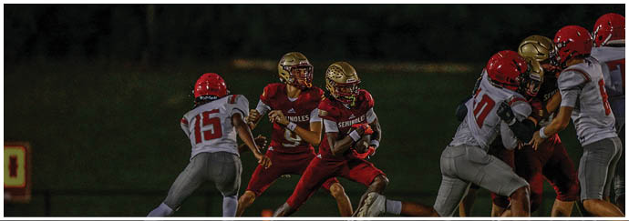 People wearing football helmets and uniforms are engaged in a play on a grassy field with some people and trees blurred in the background. Description generated by AI
