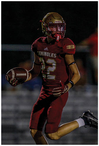 A person wearing a football uniform with a gold helmet and red pants is running with a football on a field while another person in white pants and cleats approaches. Description generated by AI