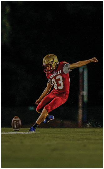 A person wearing a white and maroon football uniform with bright yellow cleats is running while holding a football and being tackled by two people in red and white uniforms on a green field. Description generated by AI