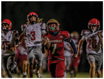 A person wearing a red sports jersey and protective helmet is crouched on a football field holding a football, preparing to snap it to a teammate. Description generated by AI
