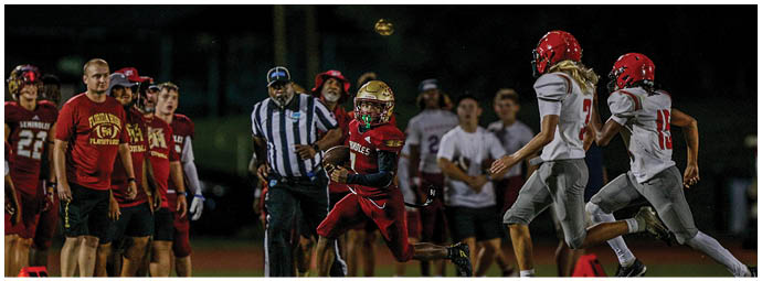 A group of people wearing football helmets and uniforms are actively playing on a field with one person holding the ball while others attempt to tackle or block. Description generated by AI