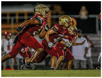 Two people in football uniforms are engaged in a play on a field, with one person in a red jersey running while holding the ball and the other in a white jersey attempting to tackle them. Description generated by AI