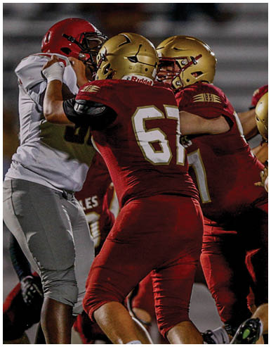 People wearing football uniforms are engaged in a tackle on a football field with blurred spectators and cheerleaders in the background. Description generated by AI