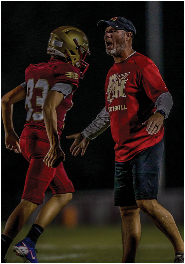 A person wearing a white and red football uniform with the number three is jumping in the air during a game while other players and cheerleaders are visible in the background. Description generated by AI