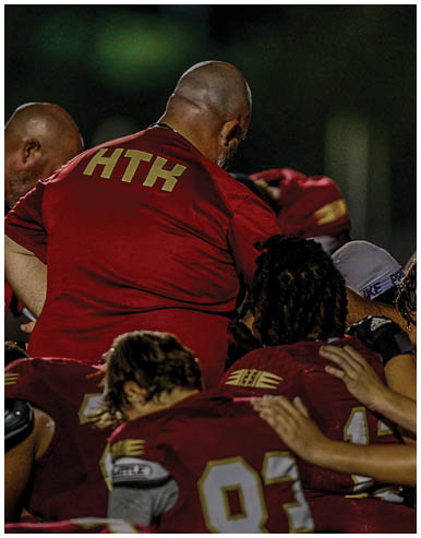 People in football uniforms are actively engaged in a play on a grassy field with one player in red holding the ball while others in red and white attempt to block or tackle. Description generated by AI