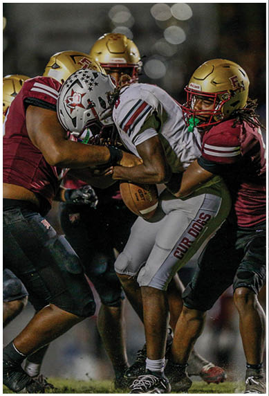 A group of people in football uniforms are actively playing on a field with one person in dark clothing tackling another person in light clothing who is holding a football. Description generated by AI