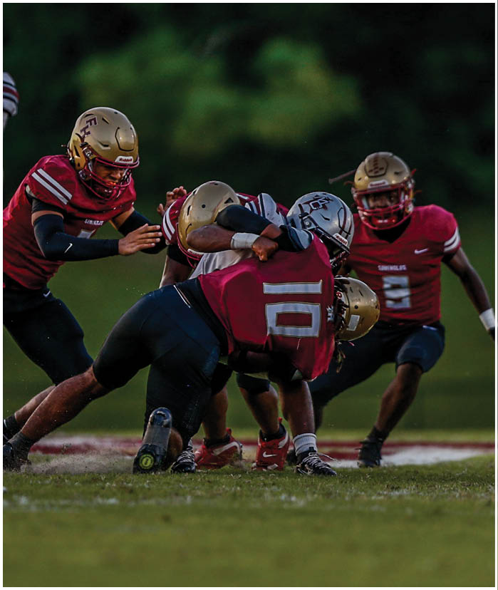 A person in a dark uniform is running with a football while being tackled by a person in a white and gold uniform during a football game. Description generated by AI