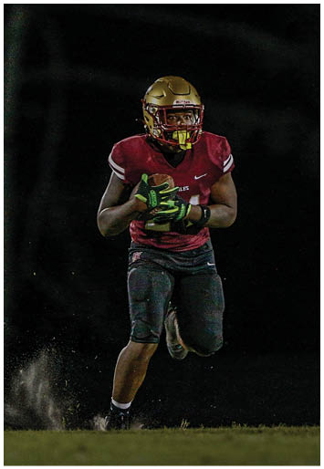 Two people wearing gold football helmets and white jerseys are engaged in a play on a dimly lit field at night. Description generated by AI