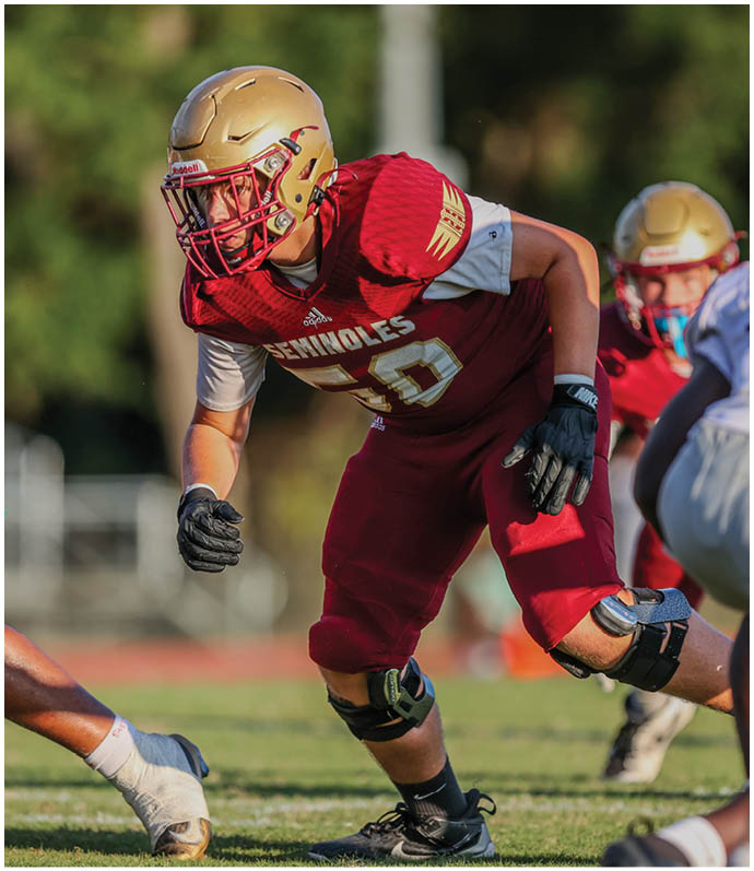 A person wearing a red football uniform with the word Seminoles and the number 50 is crouched in an athletic stance on a grassy field during a game. Description generated by AI