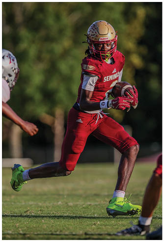 A person in red football gear and a gold helmet is running with the ball while another person in white football gear is reaching out to tackle them on a grassy field. Description generated by AI