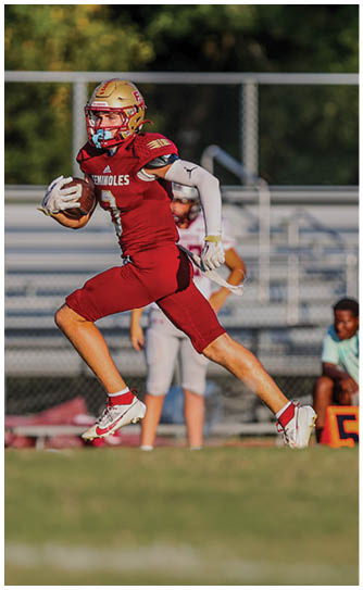A person in a red football uniform and gold helmet is running on a field while holding a football. Description generated by AI