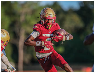 A person wearing a red football uniform and gold helmet is running while holding a football on a field with blurred trees in the background. Description generated by AI