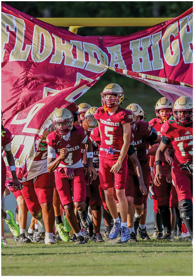 A group of people wearing maroon football uniforms and helmets run onto a field through a torn banner. Description generated by AI