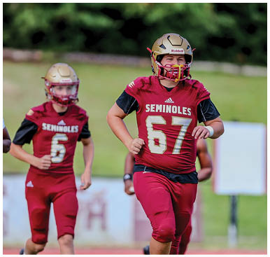 Two people wearing maroon football uniforms and gold helmets are running on a field with a blurred green background. Description generated by AI