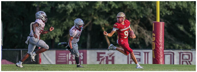 A person in a red football uniform is running with the ball while two people in white football uniforms chase them on a grassy field near a goalpost. Description generated by AI