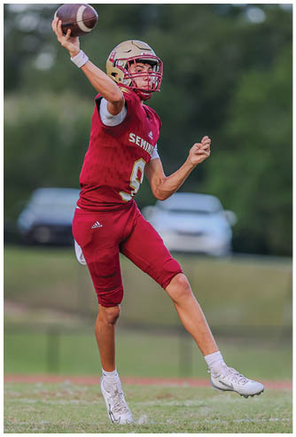 A person wearing a red football uniform and helmet is in mid-action on a grassy field with a blurred car and trees in the background. Description generated by AI