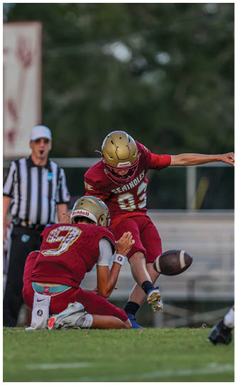 Two people in football uniforms are on a field with one holding the ball in place while the other kicks it during a game, with a referee in the background. Description generated by AI
