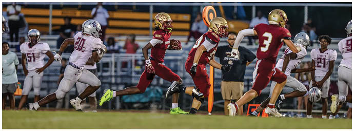 People in football uniforms are running on a field during a game with some players in red and gold and others in white and maroon. Description generated by AI