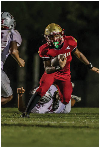 A person in a red football uniform and gold helmet is running with a football while two people in white uniforms attempt to tackle them on a grassy field at night. Description generated by AI