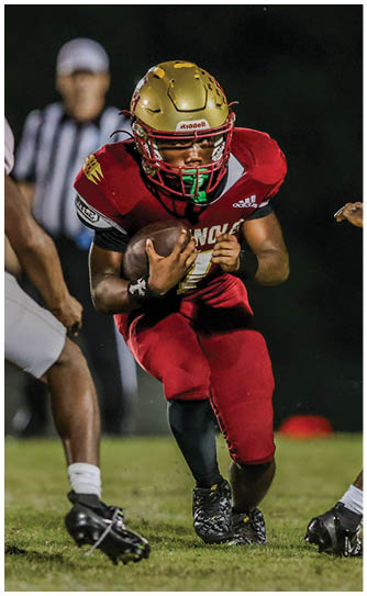 A person wearing a gold helmet and red football uniform is running while holding a football tightly during a nighttime game. Description generated by AI