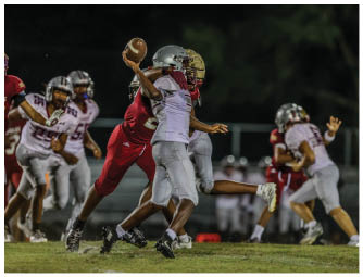 People in football uniforms are actively playing on a field at night with one person preparing to throw a football while others attempt to block or tackle. Description generated by AI