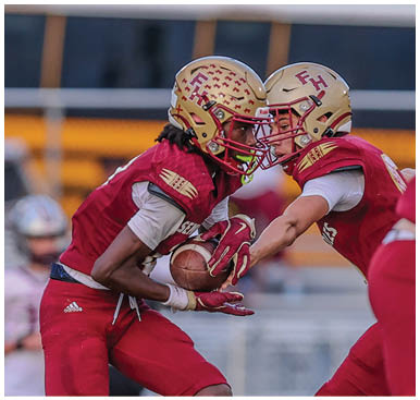 Two people in red football uniforms are exchanging a football on a grassy field during a game. Description generated by AI