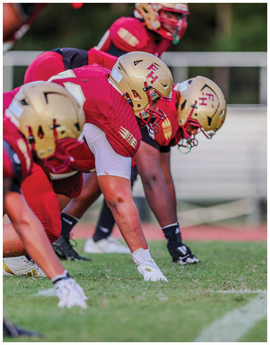 Three people in red football uniforms and gold helmets are crouched in a ready position on a grassy field during a game or practice. Description generated by AI