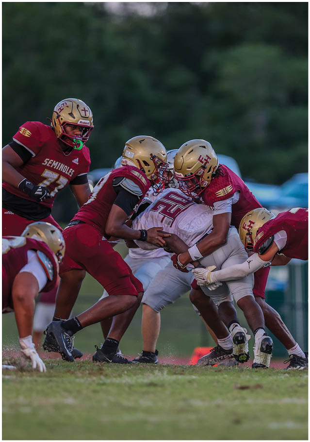 Four football players in helmets and uniforms are engaged in a tackle on a grassy field during a game. Description generated by AI