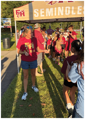 A person wearing a red shirt with “FH PTSA" printed on it stands under a tent near a sidewalk with a trash can and a "Coming Soon!" sign for a building project in the background. Description generated by AI